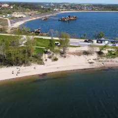 Ludzie spacerują po piaszczystej plaży nad Mietkowym zalewem. W oddali widać budynki i maszyny na wodzie.