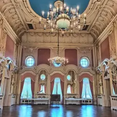 Wnętrze pałacu poznańskiego. Bogato zdobiona sala balowa z dwoma żyrandolami, okrągłymi oknami i różowymi zasłonami.