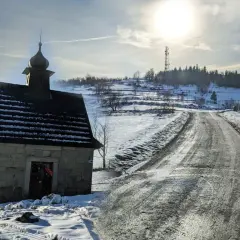Kamienna kapliczka na zboczu ośnieżonej koskowa góra, z drogą wijącą się w słońcu.