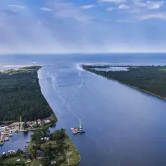Szerokie ujście Wisły do Bałtyku, otoczone lasami i niewielką osadą z portem. W oddali widać morze.