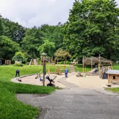 Plac zabaw w parku Kościuszki, otoczony zielenią, z piaskownicą, zjeżdżalniami i domkiem dla dzieci.