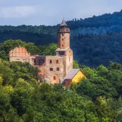 Ruiny zamku na wzgórzu otoczone zielenią, w oddali widać las i niebo. Widok przypomina krajobraz nad jeziorem Bystrzyckim.