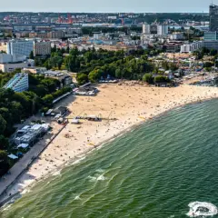 Słoneczny bulwar nadmorski Gdynia tętni życiem. Plaża pełna ludzi, a w oddali port z żaglówkami i miejska panorama.