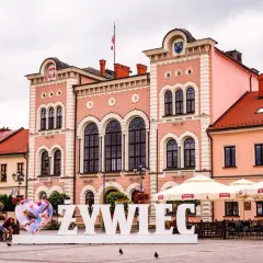 Duże białe litery "ŻYWIEC" na rynku, obok serca z ptaszkiem. W tle różowy ratusz i kawiarniane parasole.
