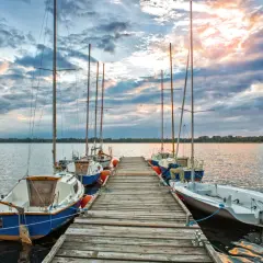 Słoneczny zachód nad pogorią jezioro. Drewniane molo z żaglówkami, chmury odbijające się w wodzie.