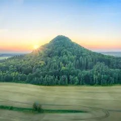 Zalesione wzgórze przypomina wygasłe wulkany w Polsce. Słońce wschodzi nad krajobrazem pól i lasów.
