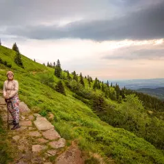 Kobieta z kijkami na kamienistej ścieżce, podziwia widok na zbocze górskie porośnięte drzewami, w oddali widać doliny. To najwyższy szczyt Sudetów.