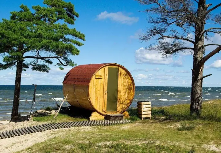Drewniana beczka-sauna na plaży, jak królowa Bałtyku, wita gości. W tle morze i niebo.