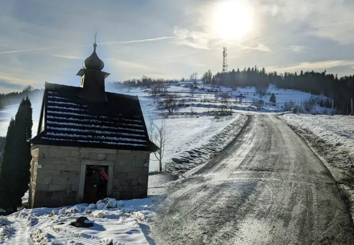 Kamienna kapliczka na zboczu ośnieżonej koskowa góra, z drogą wijącą się w słońcu.