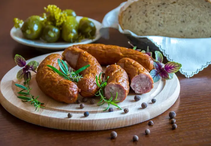Kiełbasa podwawelska na desce z ziołami, obok ogórki i chleb.
