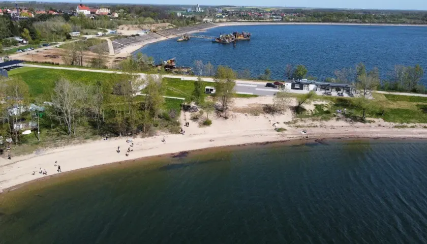Ludzie spacerują po piaszczystej plaży nad Mietkowym zalewem. W oddali widać budynki i maszyny na wodzie.