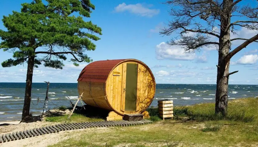 Drewniana beczka-sauna na plaży, jak królowa Bałtyku, wita gości. W tle morze i niebo.