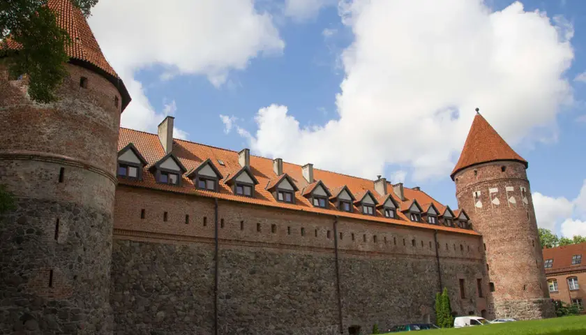 Zamek w Bytowie, z kamiennymi murami i ceglanymi wieżami, prezentuje się majestatycznie pod błękitnym niebem z chmurami.