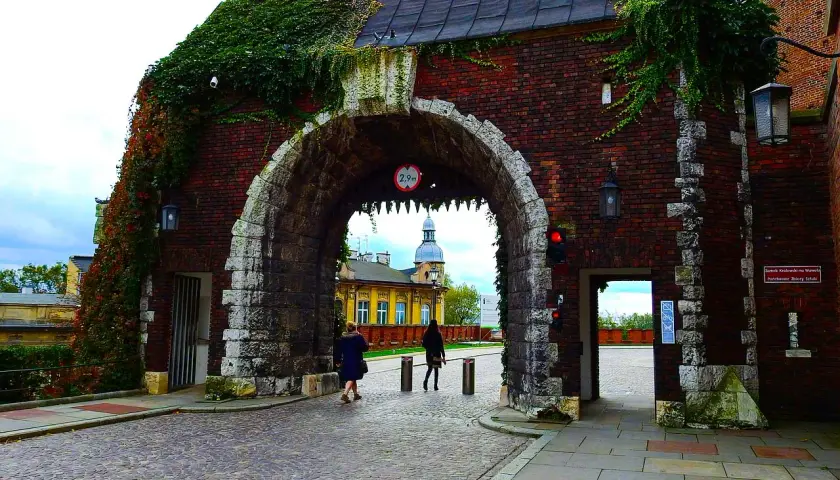 Brama wjazdowa na Wawel, porośnięta bluszczem, zaprasza do zwiedzania. Kamienna konstrukcja i ceglane mury tworzą historyczny klimat.