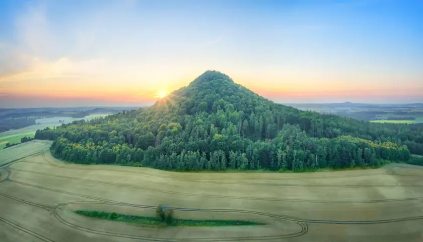 Zalesione wzgórze przypomina wygasłe wulkany w Polsce. Słońce wschodzi nad krajobrazem pól i lasów.