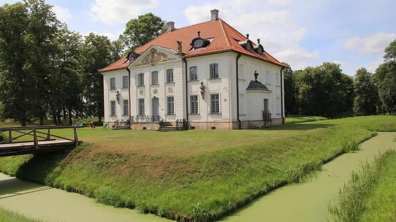 Pałac w Choroszczy otoczony fosą i zielenią, z drewnianym mostkiem prowadzącym do rezydencji.