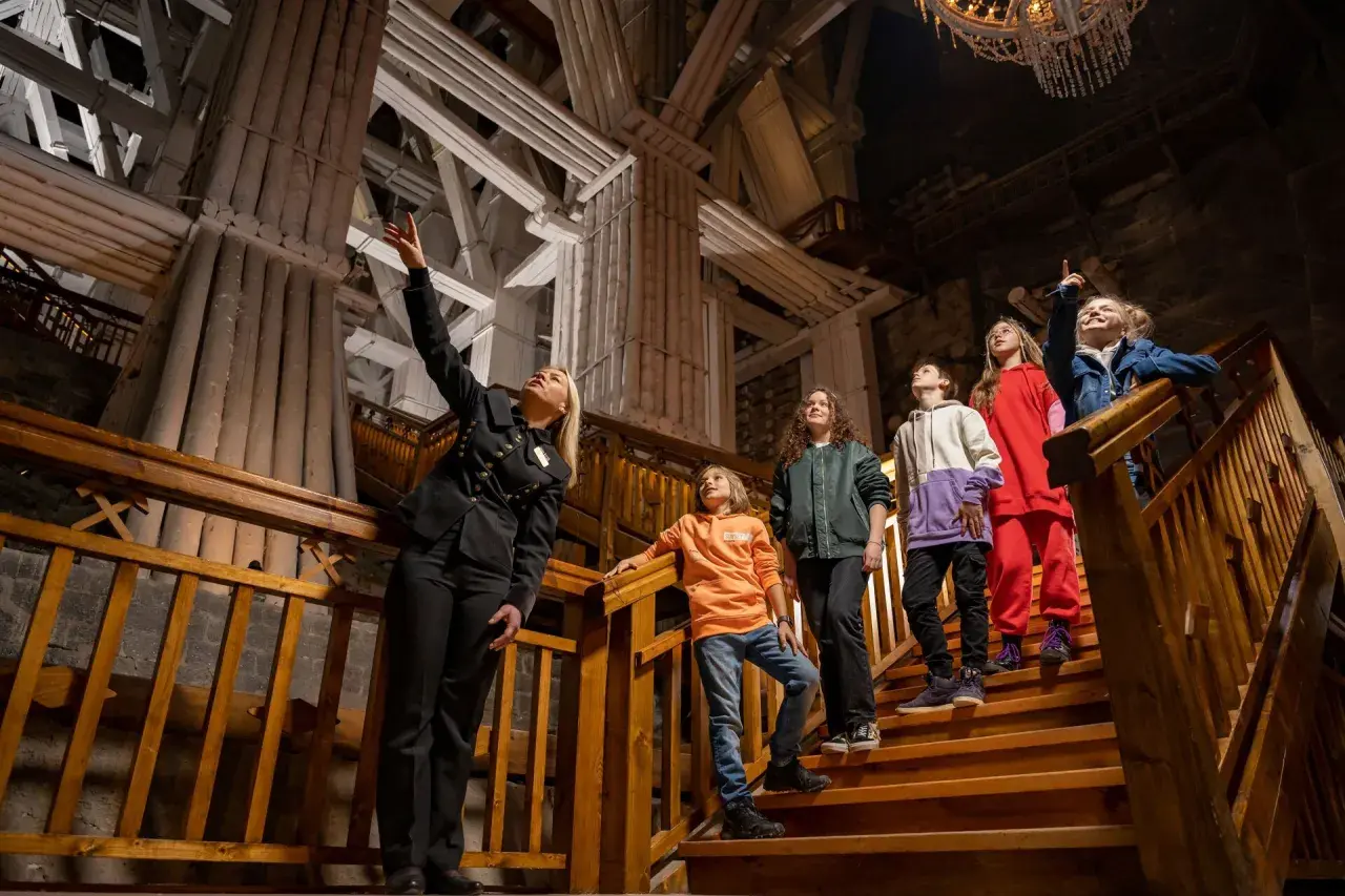 Przewodniczka oprowadza grupę dzieci po Kopalni Soli "Wieliczka". Wszyscy są ubrani na cebulkę, co sugeruje stałą, niską temperaturę w kopalni.