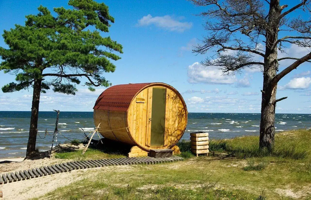 Drewniana beczka-sauna na plaży, jak królowa Bałtyku, wita gości. W tle morze i niebo.