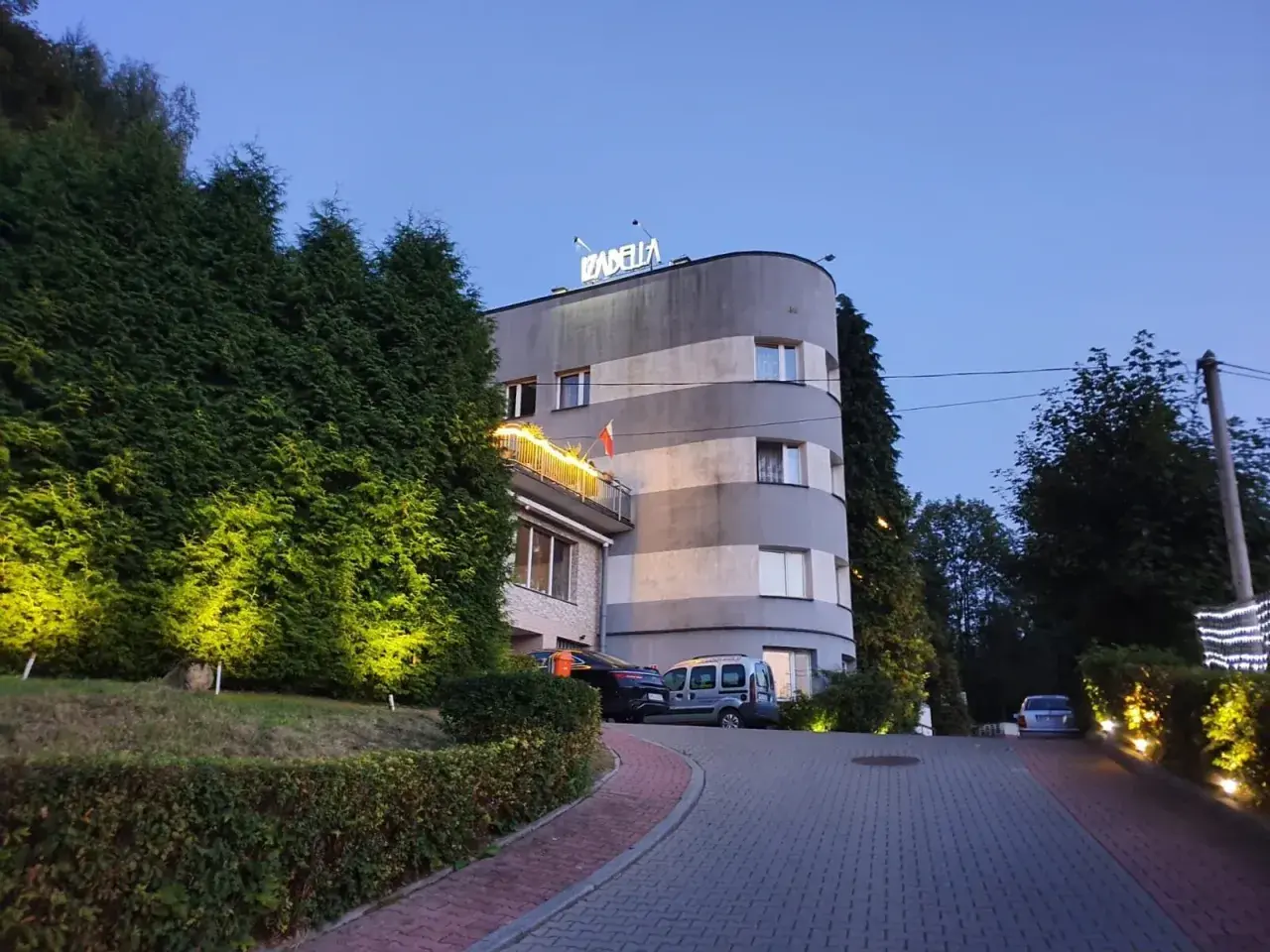 Rezydencja Izabella nocą. Elegancka budowla z podświetlanymi żywopłotami i parkingiem.