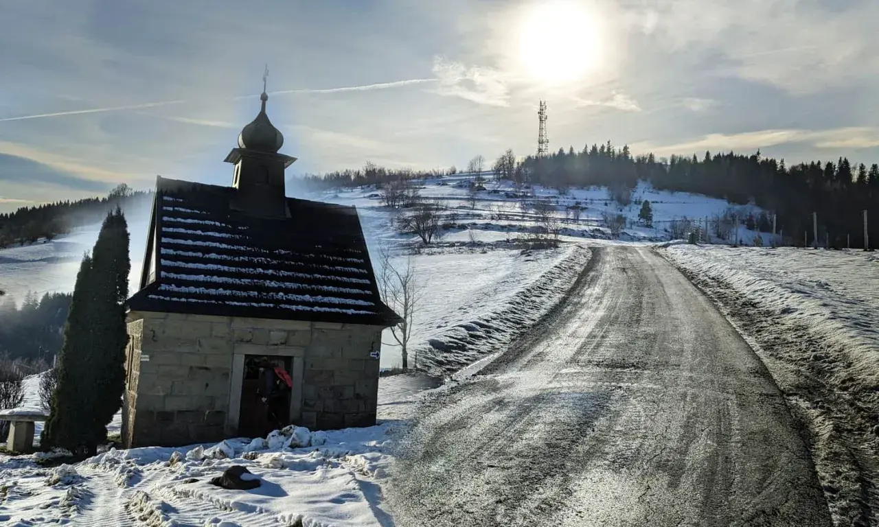 Kamienna kapliczka na zboczu ośnieżonej koskowa góra, z drogą wijącą się w słońcu.