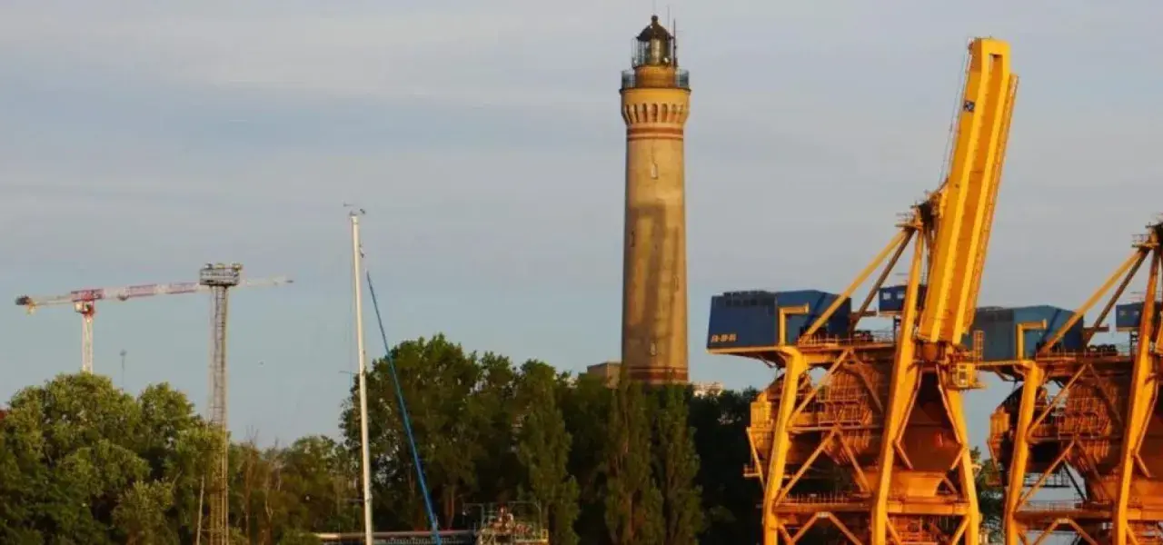 Najwyższa latarnia morska w Polsce, strzegąca portu, góruje nad żółtymi dźwigami i zielenią.