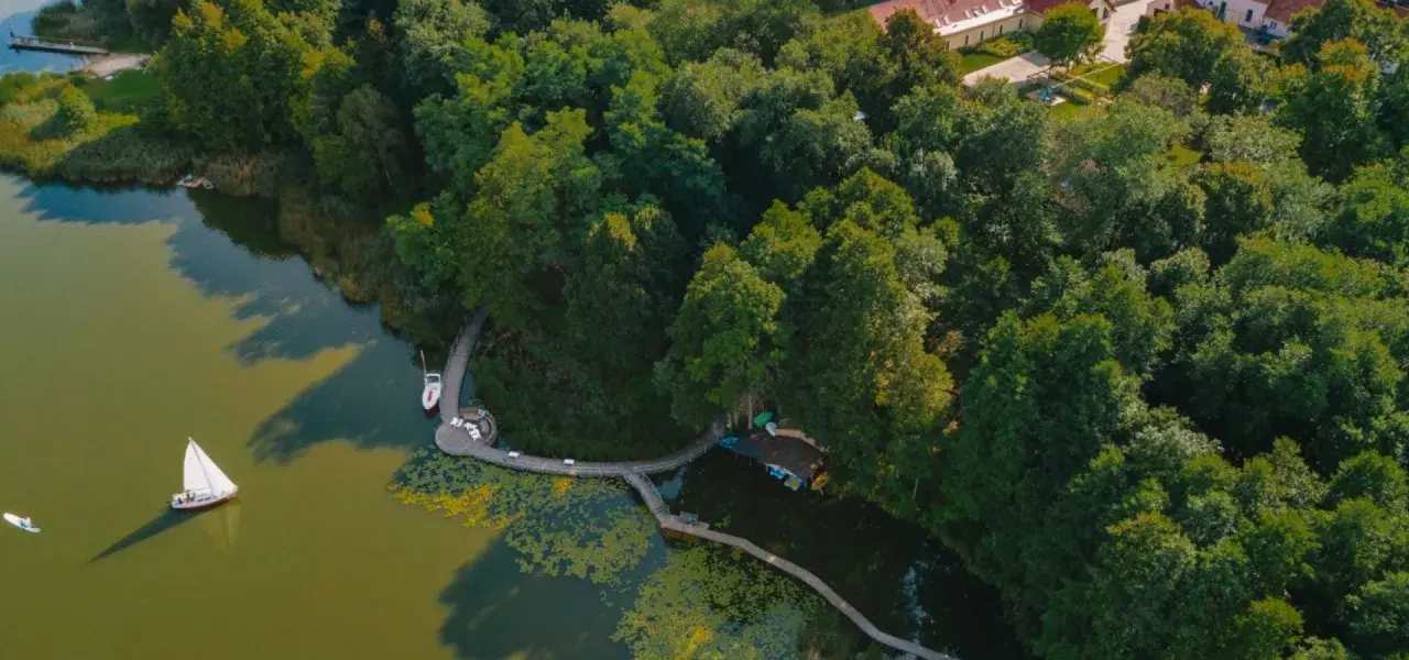 Widok z lotu ptaka na jezioro z żaglówką, drewnianą kładką i otaczającym lasem.