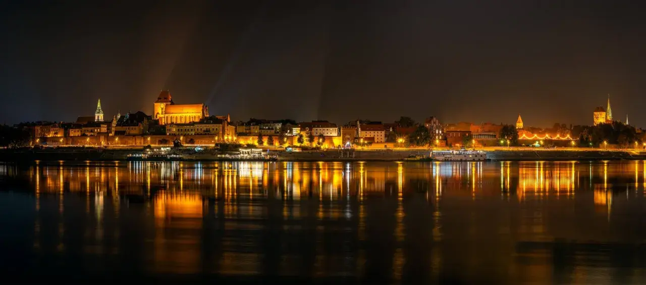 Nocny widok na oświetlony bulwar w Toruniu z odbiciem świateł w Wiśle.