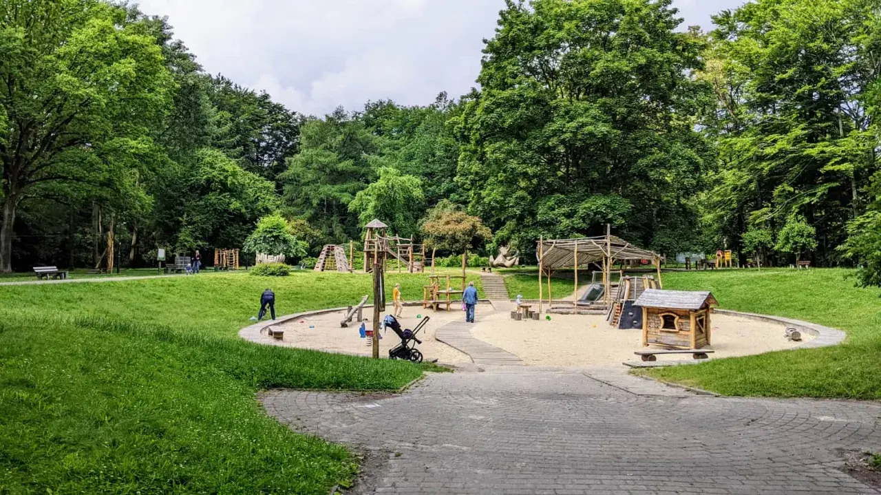 Plac zabaw w parku Kościuszki, otoczony zielenią, z piaskownicą, zjeżdżalniami i domkiem dla dzieci.