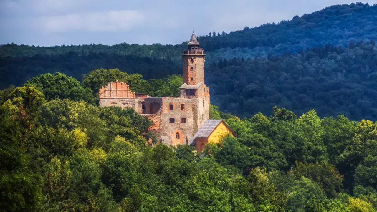 Ruiny zamku na wzgórzu otoczone zielenią, w oddali widać las i niebo. Widok przypomina krajobraz nad jeziorem Bystrzyckim.