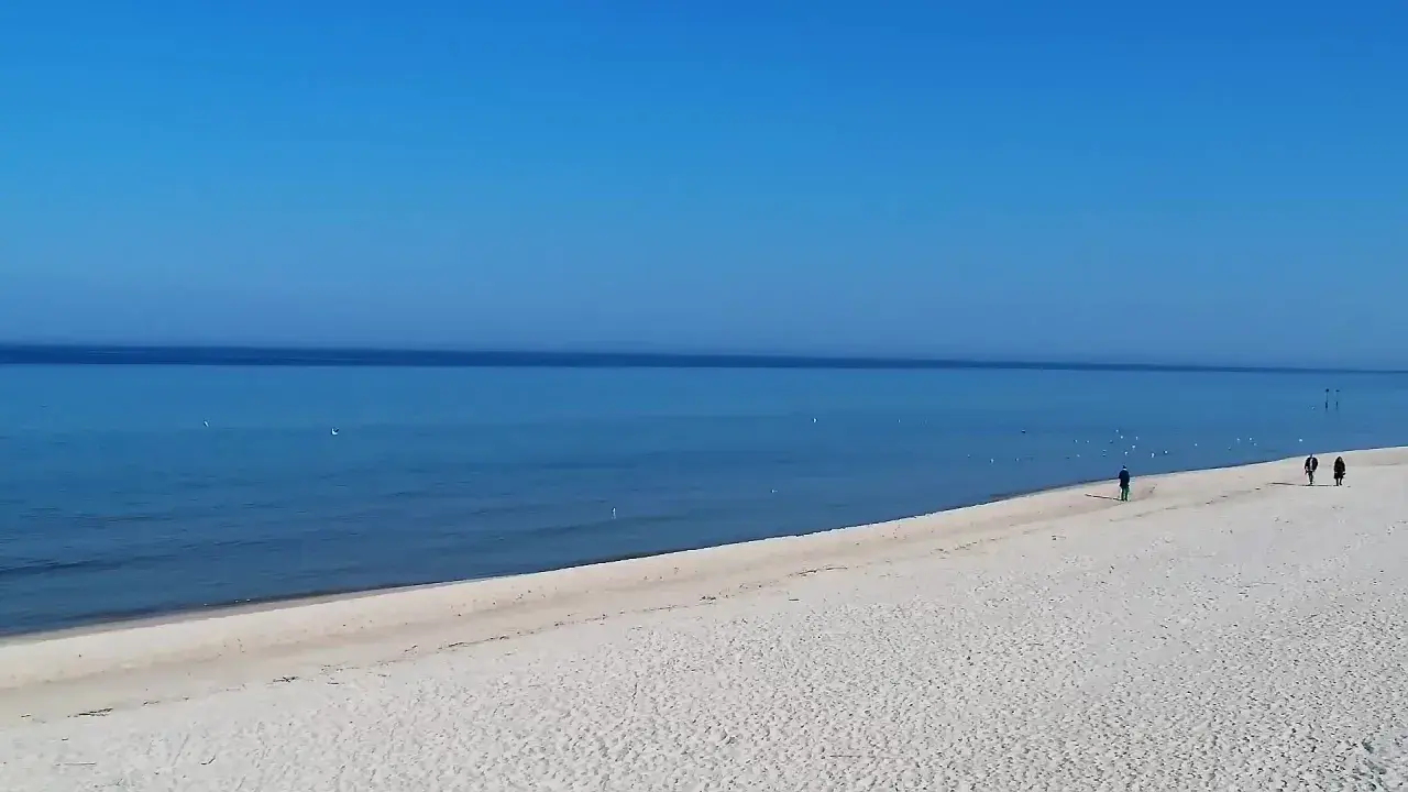 Grzybowo nadmorskie: zejście na plażę. Dwie osoby spacerują po piaszczystej plaży, morze i błękitne niebo.