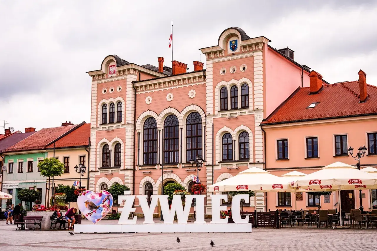 Duże białe litery "ŻYWIEC" na rynku, obok serca z ptaszkiem. W tle różowy ratusz i kawiarniane parasole.