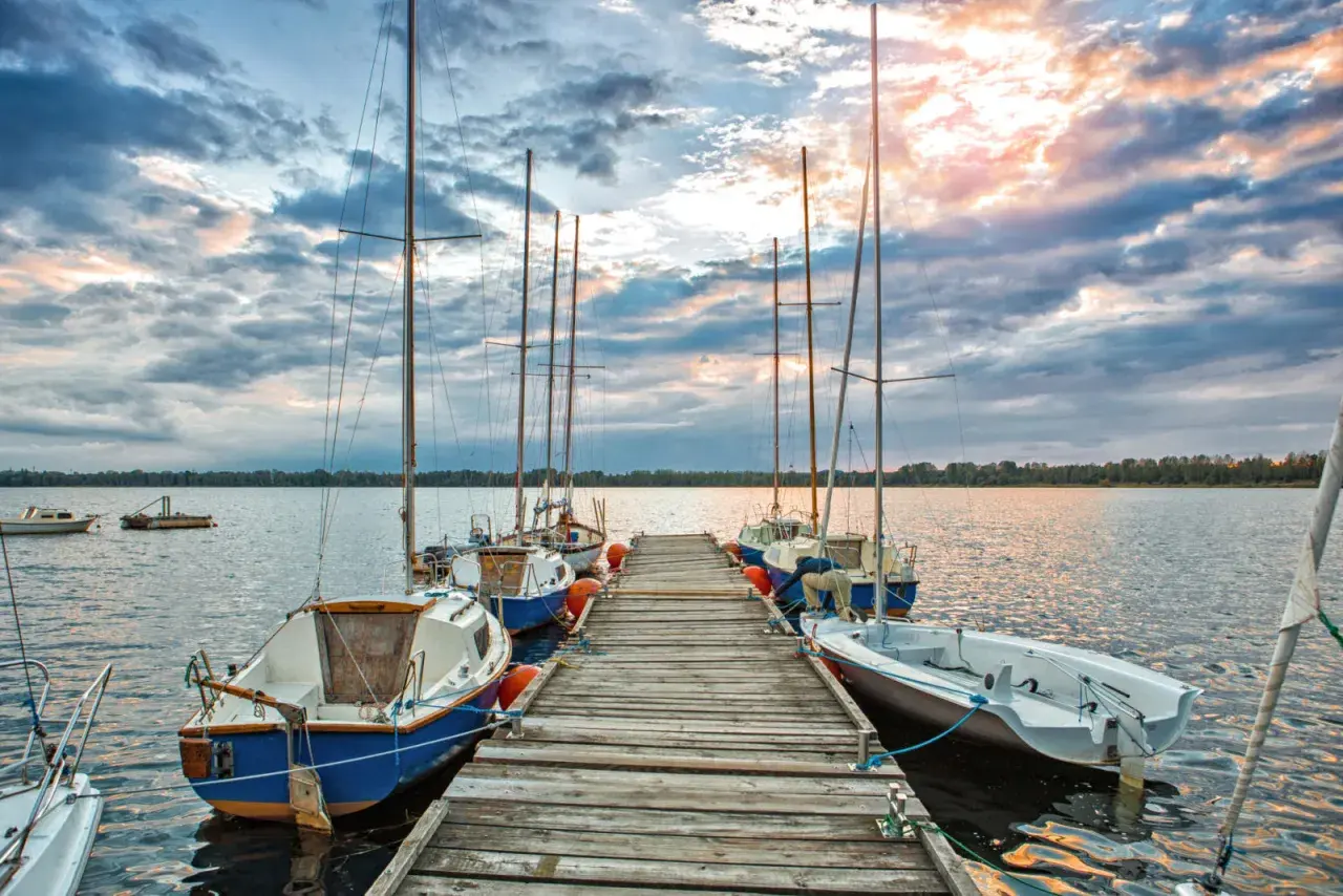Słoneczny zachód nad pogorią jezioro. Drewniane molo z żaglówkami, chmury odbijające się w wodzie.