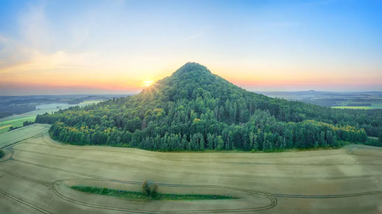 Zalesione wzgórze przypomina wygasłe wulkany w Polsce. Słońce wschodzi nad krajobrazem pól i lasów.