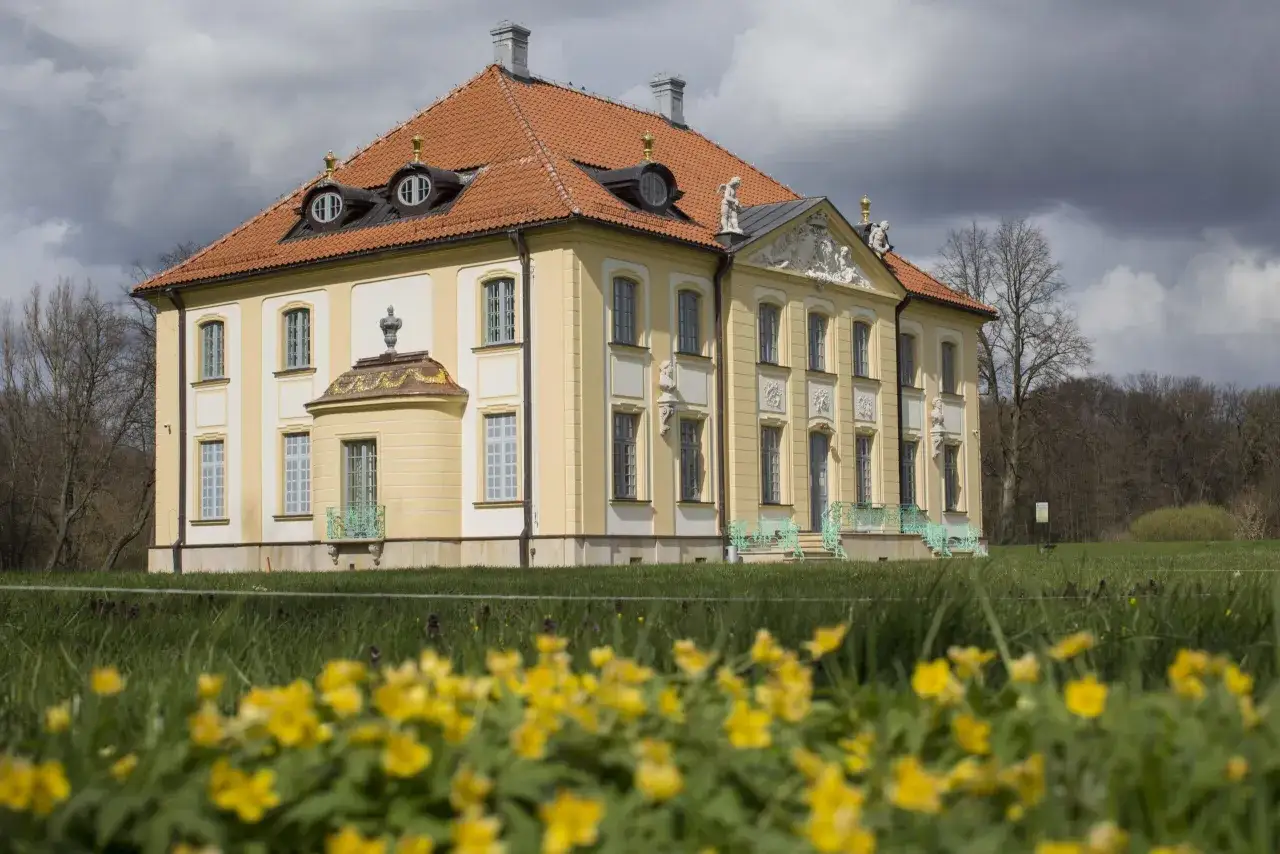 Pałac w Choroszczy, żółty, barokowy, z pomarańczowym dachem i ozdobnymi lukarnami, otoczony zielenią i kwitnącymi wiosną kwiatami.