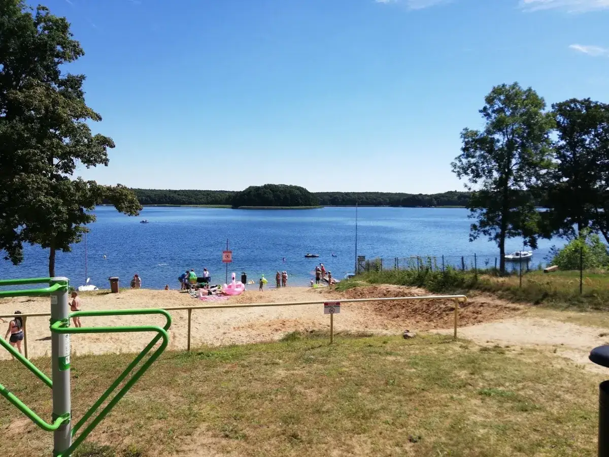 Słoneczny dzień nad jeziorem Niesłysz. Ludzie odpoczywają na piaszczystej plaży, niektórzy kąpią się w błękitnej wodzie.