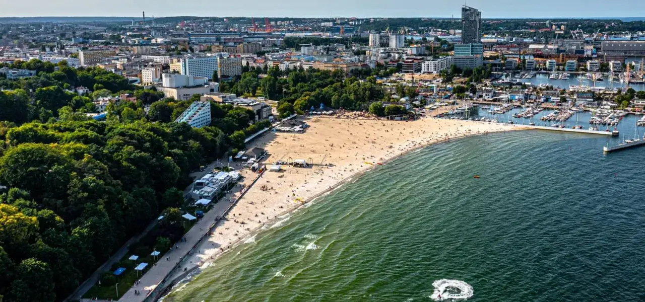 Słoneczny bulwar nadmorski Gdynia tętni życiem. Plaża pełna ludzi, morze i marina jachtowa tworzą malowniczy krajobraz.