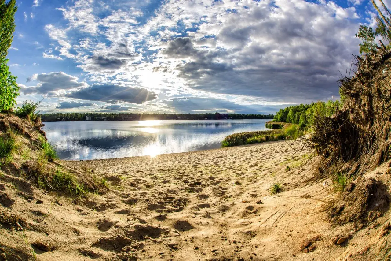 Piaskowa plaża nad jeziorem, zarośla i las w tle. Słońce przebija się przez chmury, tworząc refleksy na wodzie. To idealne miejsce na odpoczynek nad pogorią jezioro.