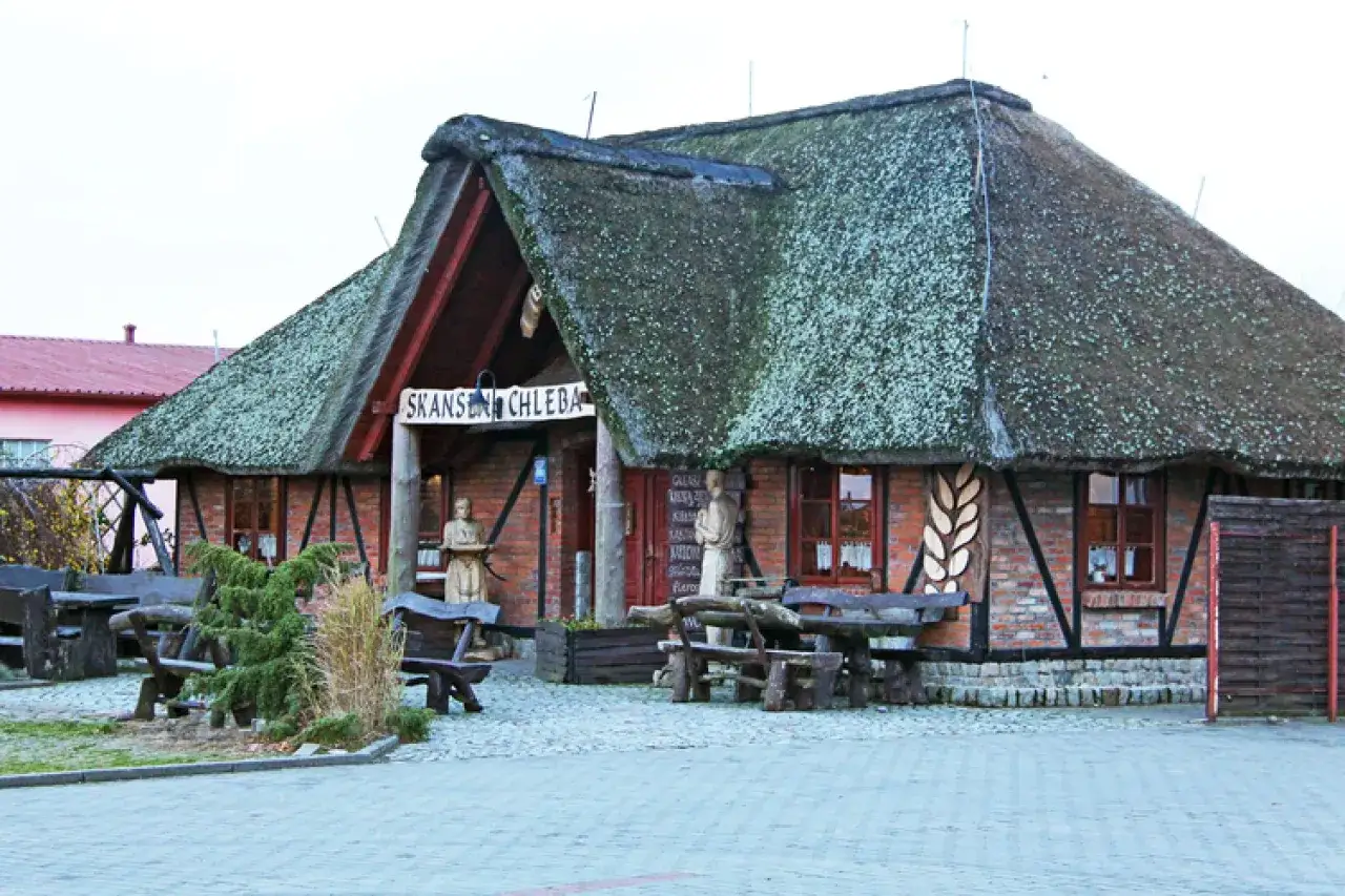 Skansen Chleba Ustronie Morskie: cennik atrakcji i wypieków. Drewniane ławy i stoły przed budynkiem z krytym strzechą dachem.