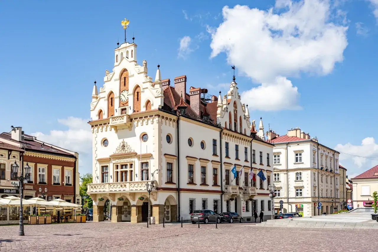 Rzeszów rynek: ratusz z zegarem i złotym orłem, kamienice, kawiarniane stoliki pod parasolami, samochody i błękitne niebo z chmurami.