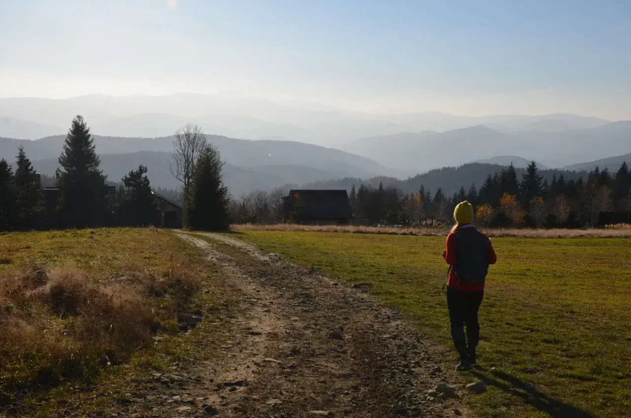 Widok na pasmo górskie z chatką i turystką na ścieżce. Koskowa Góra w oddali.