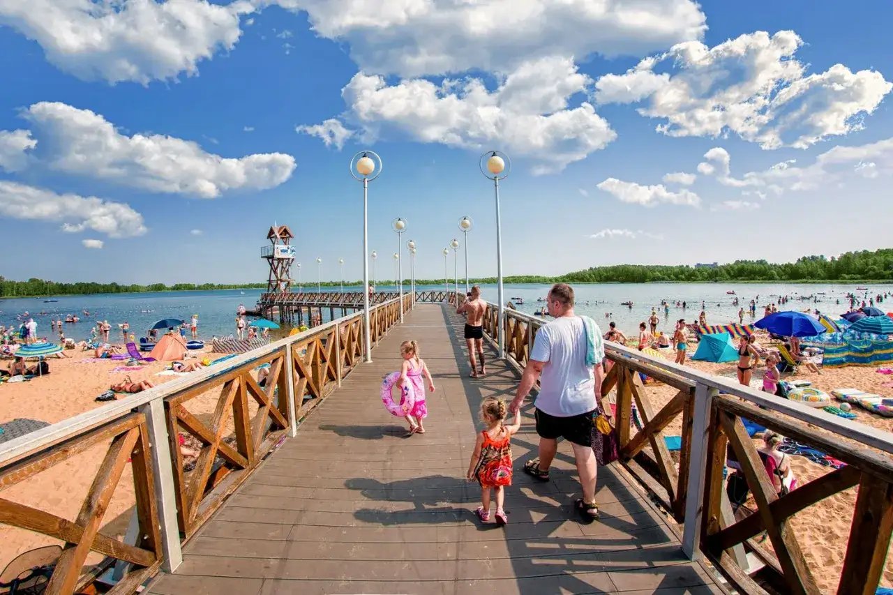 Rodzina spaceruje po molo na plaży nad zbiornikiem Pogoria III. Słoneczny dzień, dużo ludzi odpoczywa.