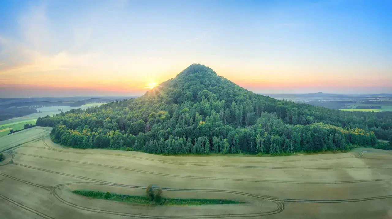 Wschód słońca nad zalesionym wzgórzem, które jest częścią szlaku wygasłych wulkanów. Pola uprawne otaczają zielony stożek.