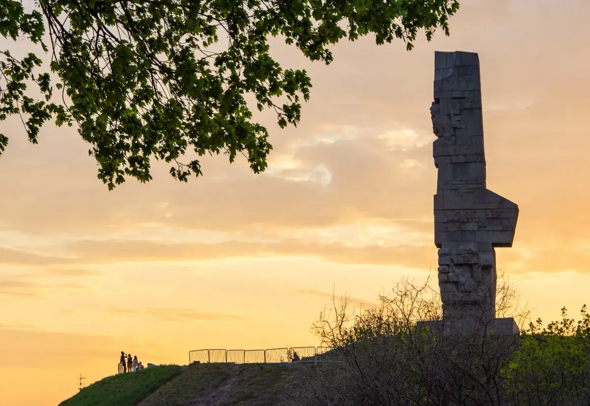 Wieczorne zwiedzanie Westerplatte. Monumentalny pomnik na tle zachodzącego słońca, z grupą turystów na wzgórzu.